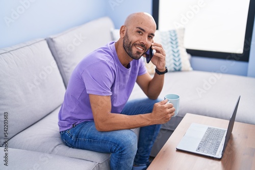 Young bald man talking on smartphone drinking coffee at home