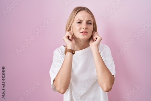 Young caucasian woman standing over pink background covering ears with fingers with annoyed expression for the noise of loud music. deaf concept.