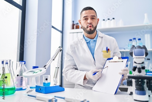 Hispanic man working at scientist laboratory holding blank clipboard making fish face with mouth and squinting eyes  crazy and comical.