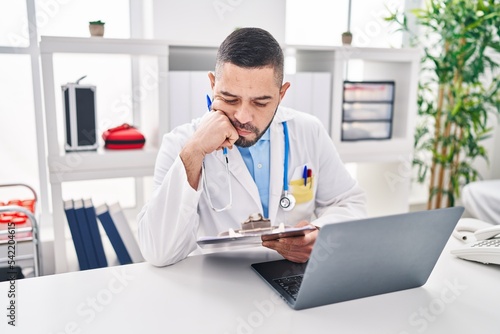 Young latin man doctor using laptop reading medical report at clinic