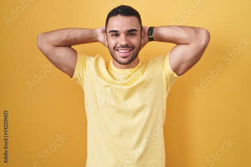Young hispanic man standing over yellow background relaxing and stretching, arms and hands behind head and neck smiling happy