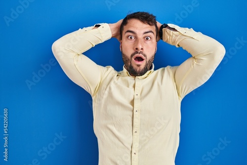 Handsome hispanic man standing over blue background crazy and scared with hands on head, afraid and surprised of shock with open mouth © Krakenimages.com