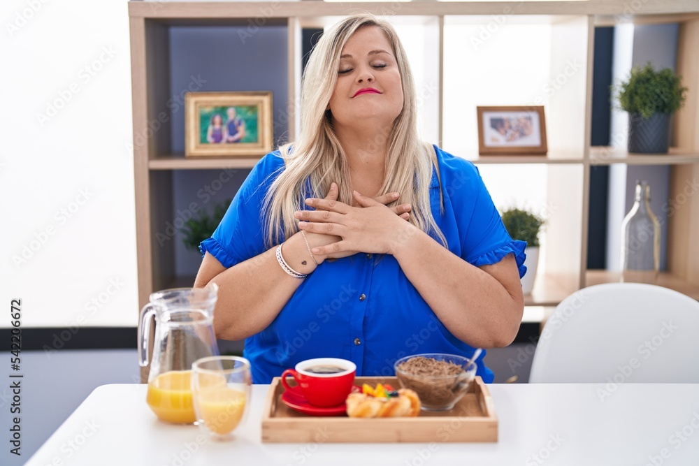 Caucasian plus size woman eating breakfast at home smiling with hands on chest with closed eyes and grateful gesture on face. health concept. Caucasian plus size woman eating breakfast at home smiling with hands on chest with closed eyes and grateful gesture on face. health concept.