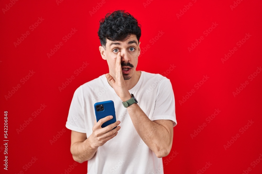 Hispanic man using smartphone over red background hand on mouth telling secret rumor, whispering malicious talk conversation Hispanic man using smartphone over red background hand on mouth telling secret rumor, whispering malicious talk conversation