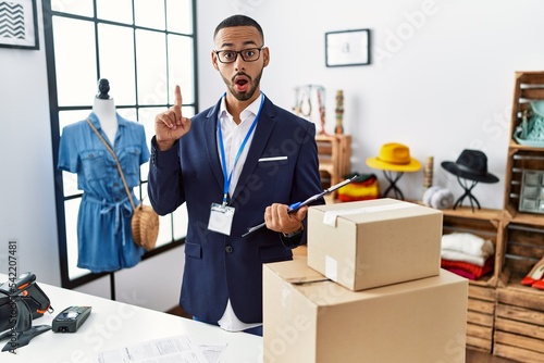 African american man working as manager at retail boutique amazed and surprised looking up and pointing with fingers and raised arms.