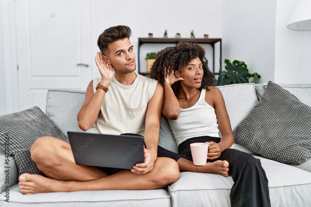 Young interracial couple using laptop at home sitting on the sofa smiling with hand over ear listening an hearing to rumor or gossip. deafness concept. Young interracial couple using laptop at home sitting on the sofa smiling with hand over ear listening an hearing to rumor or gossip. deafness concept.
