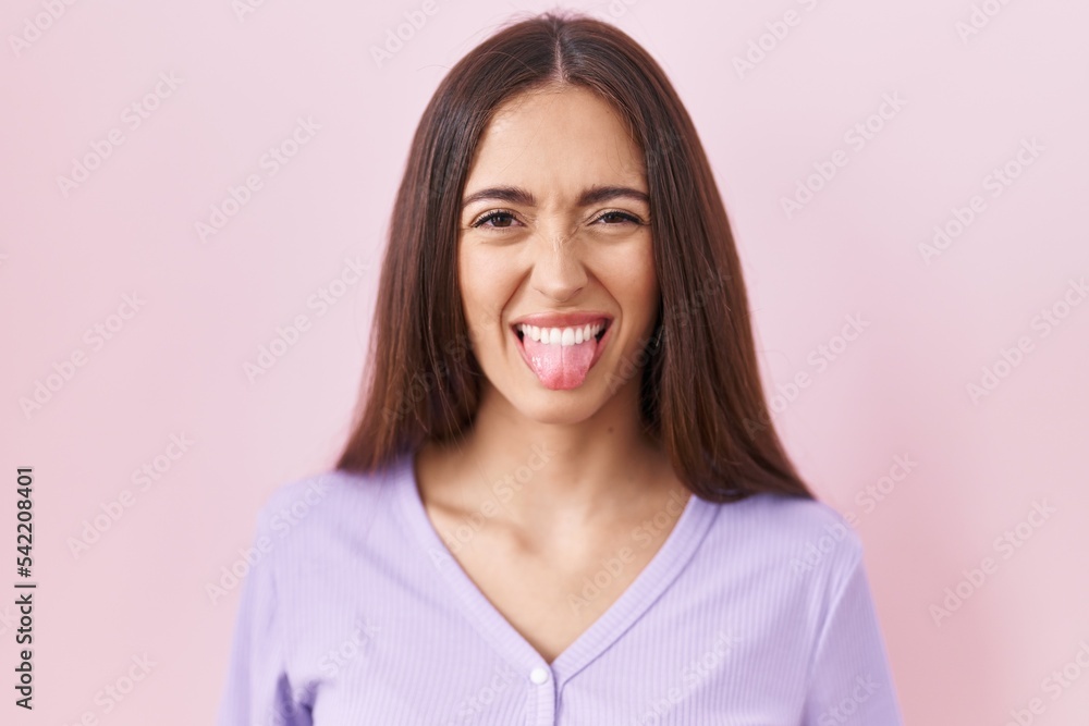 Young hispanic woman with long hair standing over pink background sticking tongue out happy with funny expression. emotion concept. Young hispanic woman with long hair standing over pink background sticking tongue out happy with funny expression. emotion concept.