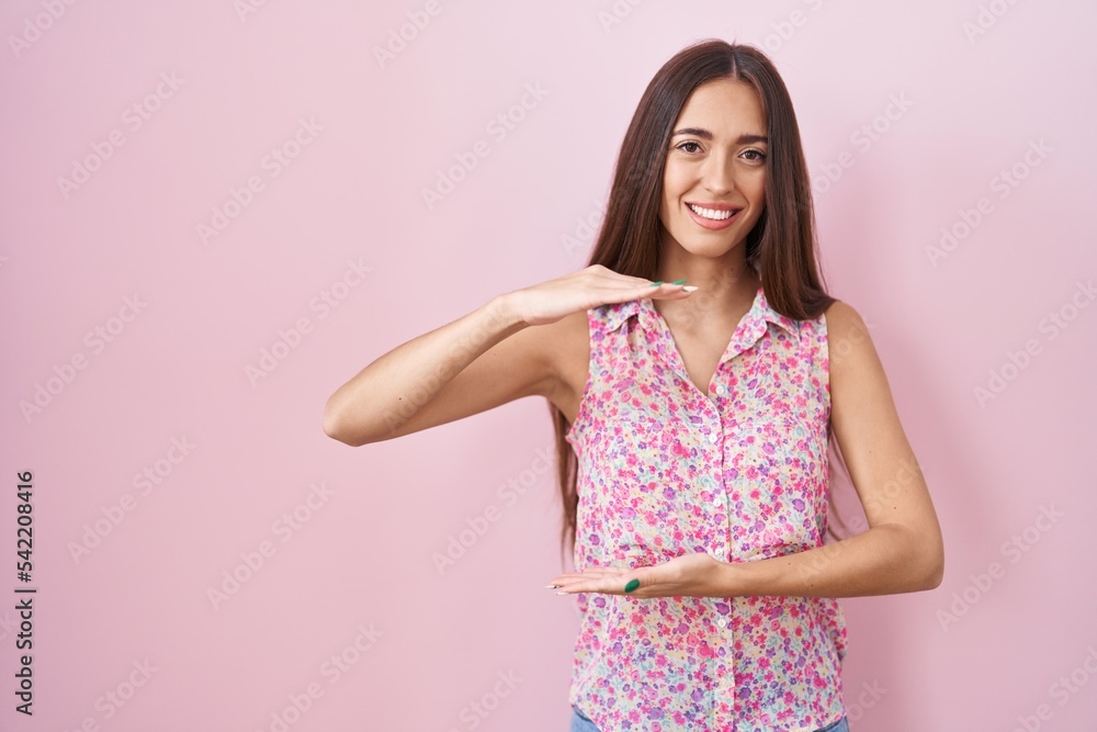 Young hispanic woman with long hair standing over pink background gesturing with hands showing big and large size sign, measure symbol. smiling looking at the camera. measuring concept.