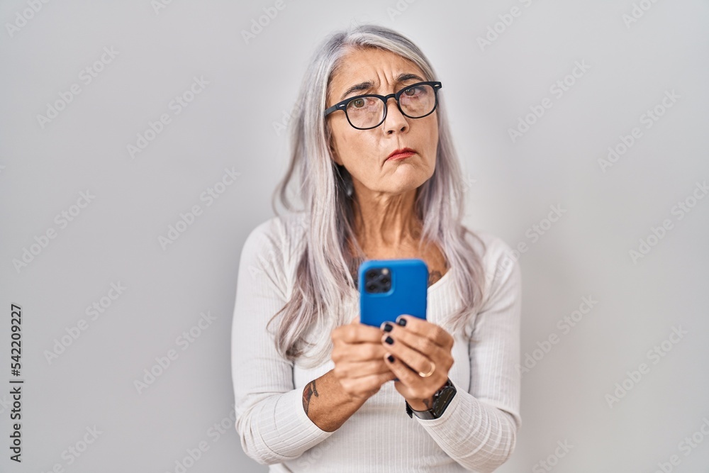 Middle age woman with grey hair using smartphone typing message clueless and confused expression. doubt concept.