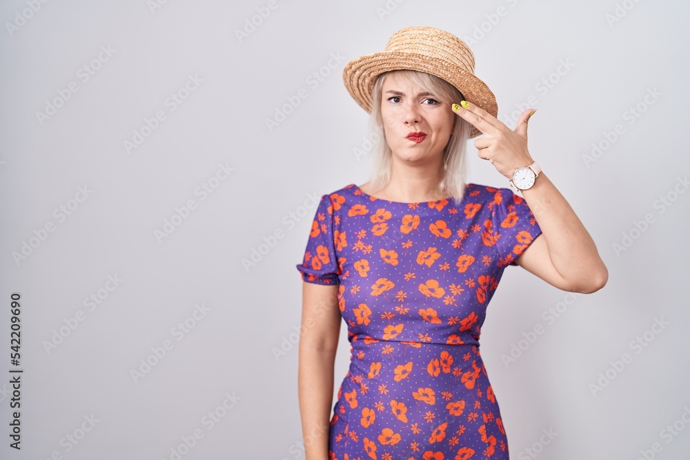 Young caucasian woman wearing flowers dress and summer hat shooting and killing oneself pointing hand and fingers to head like gun, suicide gesture. Young caucasian woman wearing flowers dress and summer hat shooting and killing oneself pointing hand and fingers to head like gun, suicide gesture.
