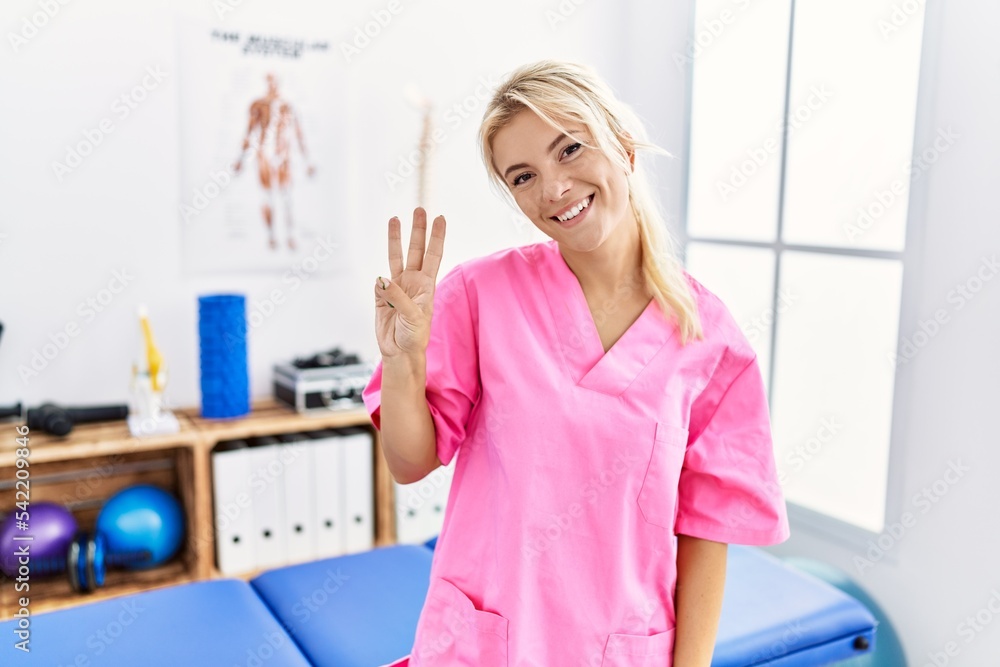 Young caucasian woman working at pain recovery clinic showing and pointing up with fingers number three while smiling confident and happy. Young caucasian woman working at pain recovery clinic showing and pointing up with fingers number three while smiling confident and happy.