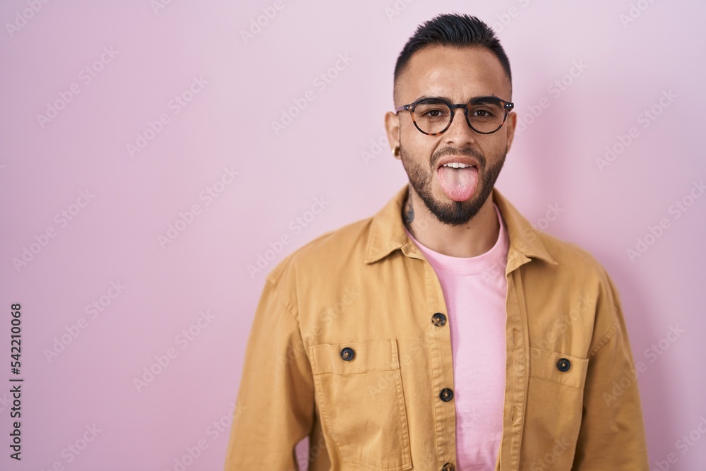 Young hispanic man standing over pink background sticking tongue out happy with funny expression. emotion concept. Young hispanic man standing over pink background sticking tongue out happy with funny expression. emotion concept.