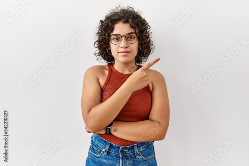 Young hispanic woman wearing glasses standing over isolated background pointing with hand finger to the side showing advertisement, serious and calm face
