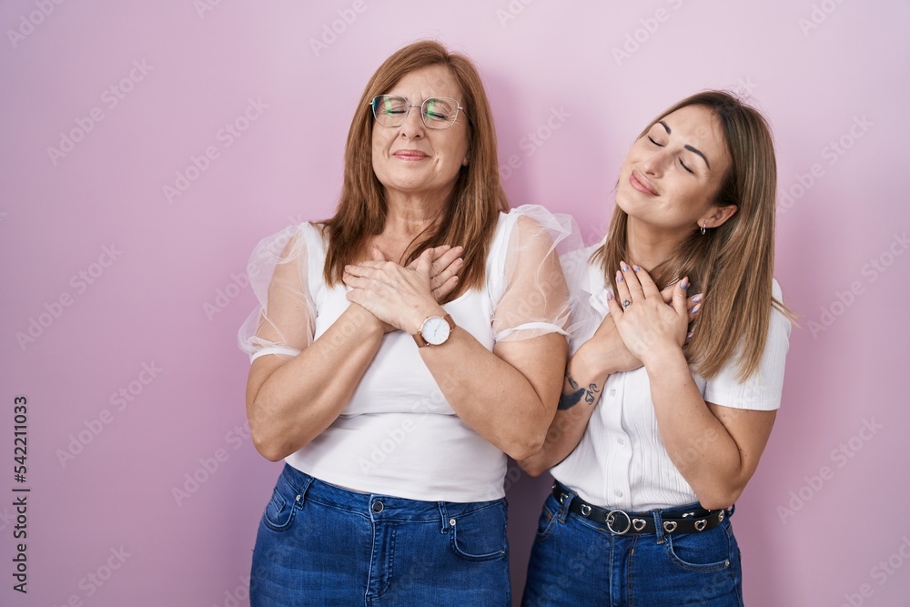 Hispanic mother and daughter wearing casual white t shirt over pink background smiling with hands on chest with closed eyes and grateful gesture on face. health concept. Hispanic mother and daughter wearing casual white t shirt over pink background smiling with hands on chest with closed eyes and grateful gesture on face. health concept.