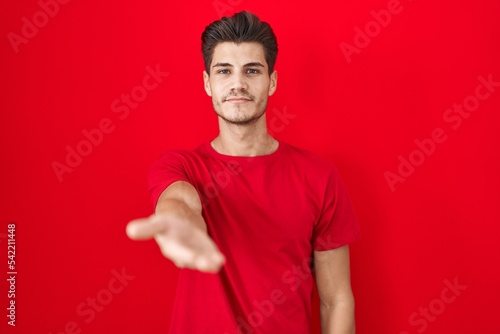 Young hispanic man standing over red background smiling cheerful offering palm hand giving assistance and acceptance.