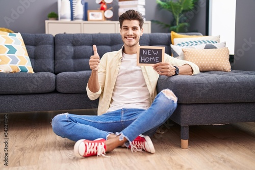 Young hispanic man holding blackboard with new home text smiling happy and positive, thumb up doing excellent and approval sign