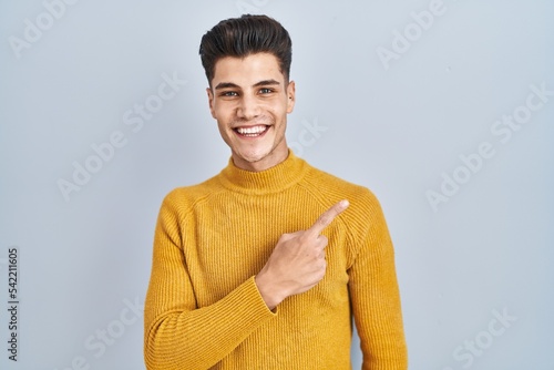 Young hispanic man standing over blue background cheerful with a smile on face pointing with hand and finger up to the side with happy and natural expression