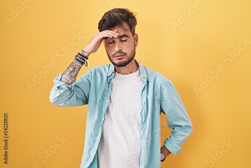 Young hispanic man with tattoos standing over yellow background worried and stressed about a problem with hand on forehead  nervous and anxious for crisis