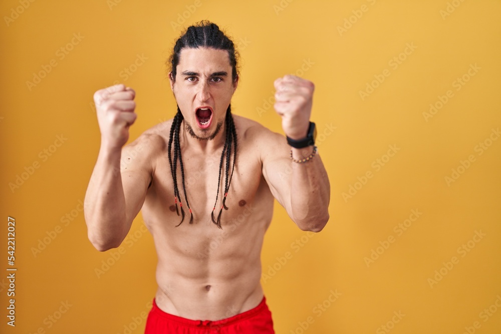 Fototapeta premium Hispanic man with long hair standing shirtless over yellow background angry and mad raising fists frustrated and furious while shouting with anger. rage and aggressive concept.