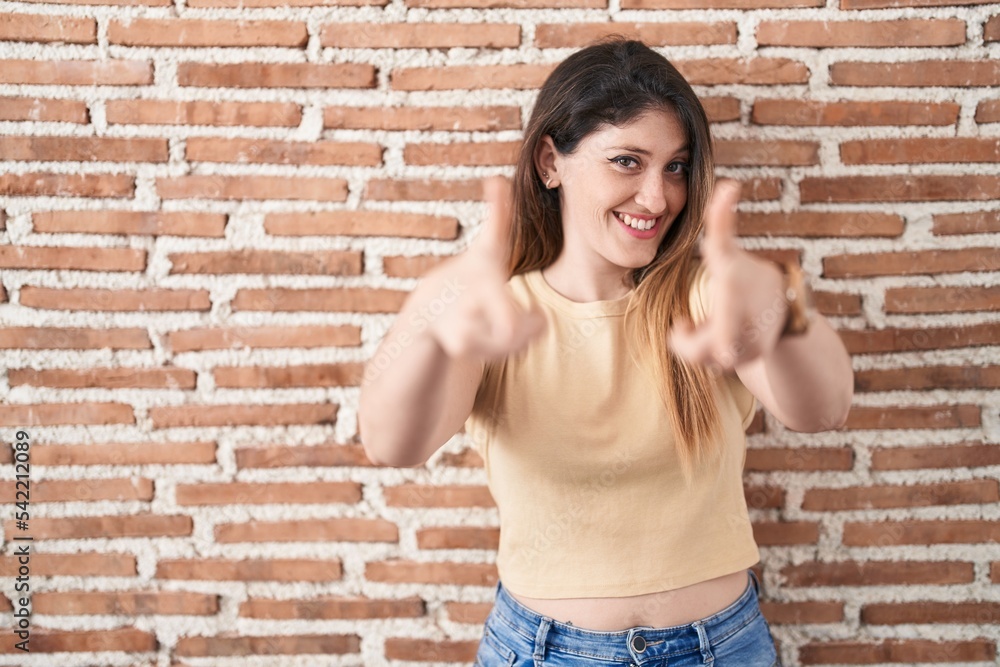 Young brunette woman standing over bricks wall pointing fingers to camera with happy and funny face. good energy and vibes. Young brunette woman standing over bricks wall pointing fingers to camera with happy and funny face. good energy and vibes.