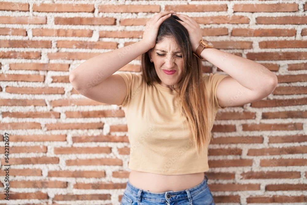 Young brunette woman standing over bricks wall suffering from headache desperate and stressed because pain and migraine. hands on head. Young brunette woman standing over bricks wall suffering from headache desperate and stressed because pain and migraine. hands on head.