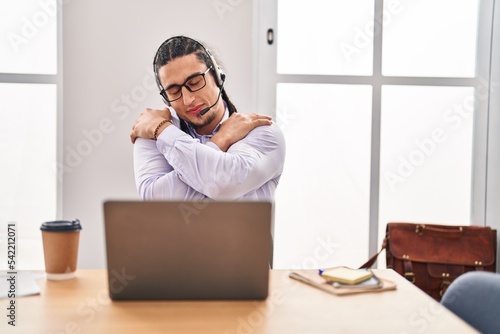 Hispanic man with long hair working using computer laptop hugging oneself happy and positive, smiling confident. self love and self care