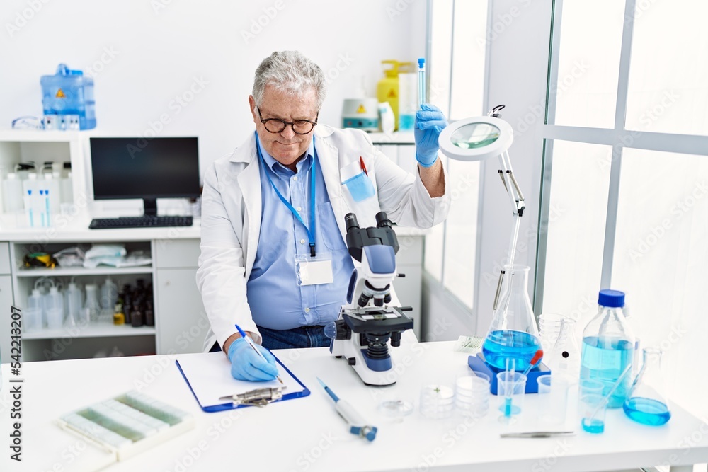Middle age grey-haired man wearing scientist uniform holding test tube and writing on clipboard at laboratory Middle age grey-haired man wearing scientist uniform holding test tube and writing on clipboard at laboratory