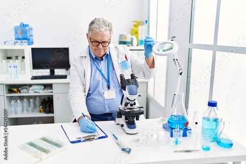 Middle age grey-haired man wearing scientist uniform holding test tube and writing on clipboard at laboratory