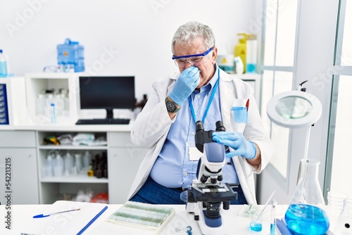 Senior caucasian man working at scientist laboratory smelling something stinky and disgusting  intolerable smell  holding breath with fingers on nose. bad smell