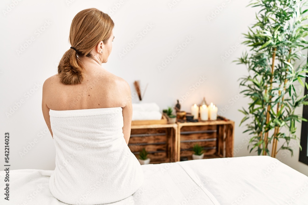 Middle age caucasian woman on back view wearing towel sitting on massage board at beauty center Middle age caucasian woman on back view wearing towel sitting on massage board at beauty center