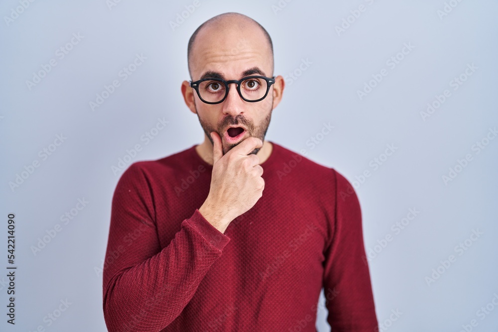 Young bald man with beard standing over white background wearing glasses looking fascinated with disbelief, surprise and amazed expression with hands on chin Young bald man with beard standing over white background wearing glasses looking fascinated with disbelief, surprise and amazed expression with hands on chin