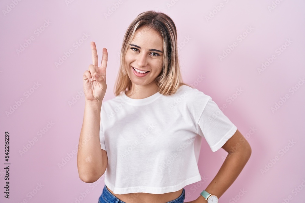 Young blonde woman standing over pink background smiling looking to the camera showing fingers doing victory sign. number two. Young blonde woman standing over pink background smiling looking to the camera showing fingers doing victory sign. number two.