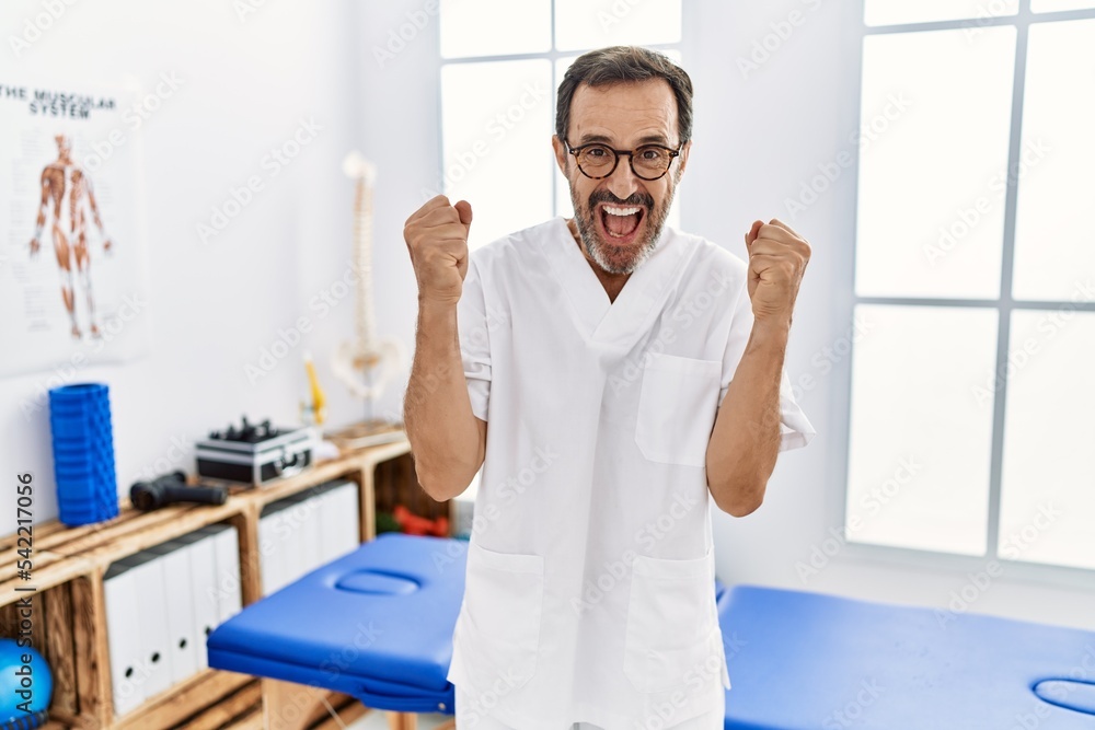 Middle age man with beard working at pain recovery clinic celebrating surprised and amazed for success with arms raised and open eyes. winner concept. Middle age man with beard working at pain recovery clinic celebrating surprised and amazed for success with arms raised and open eyes. winner concept.