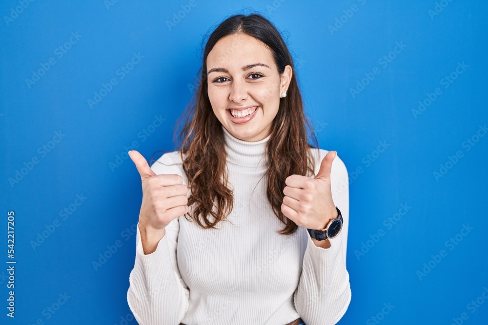 Young hispanic woman standing over blue background success sign doing positive gesture with hand, thumbs up smiling and happy. cheerful expression and winner gesture. Young hispanic woman standing over blue background success sign doing positive gesture with hand, thumbs up smiling and happy. cheerful expression and winner gesture.