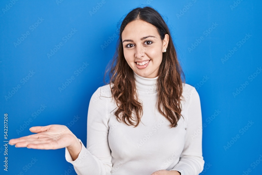 Young hispanic woman standing over blue background smiling cheerful presenting and pointing with palm of hand looking at the camera. Young hispanic woman standing over blue background smiling cheerful presenting and pointing with palm of hand looking at the camera.