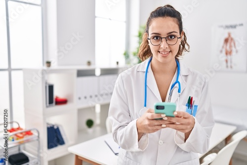 Young beautiful hispanic woman doctor smiling confident using smartphone at clinic