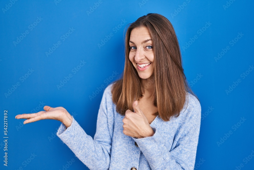 Young woman standing over blue background showing palm hand and doing ok gesture with thumbs up, smiling happy and cheerful Young woman standing over blue background showing palm hand and doing ok gesture with thumbs up, smiling happy and cheerful