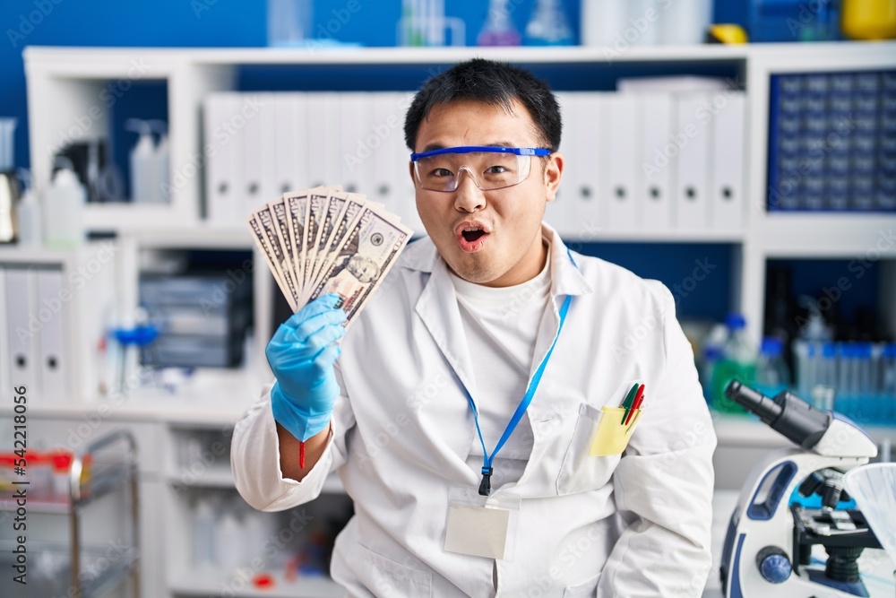 Young chinese man working at scientist laboratory holding money scared and amazed with open mouth for surprise, disbelief face Young chinese man working at scientist laboratory holding money scared and amazed with open mouth for surprise, disbelief face
