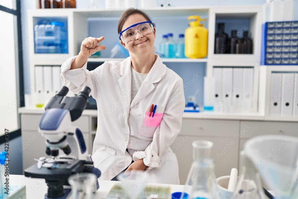 Hispanic girl with down syndrome working at scientist laboratory smiling and confident gesturing with hand doing small size sign with fingers looking and the camera. measure concept. Hispanic girl with down syndrome working at scientist laboratory smiling and confident gesturing with hand doing small size sign with fingers looking and the camera. measure concept.