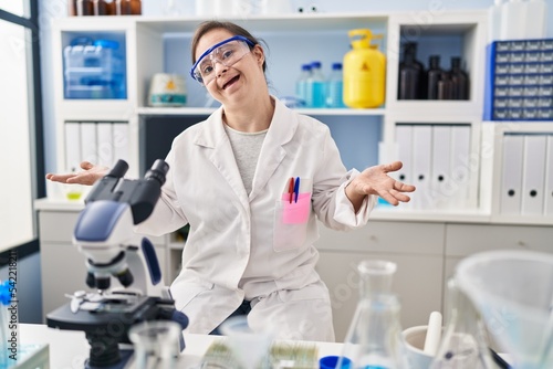 Hispanic girl with down syndrome working at scientist laboratory smiling cheerful with open arms as friendly welcome  positive and confident greetings