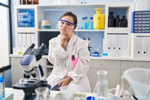 Hispanic girl with down syndrome working at scientist laboratory thinking concentrated about doubt with finger on chin and looking up wondering