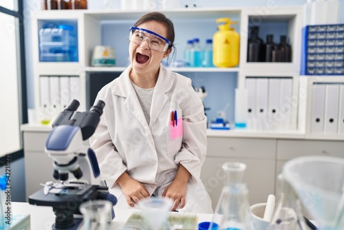 Hispanic girl with down syndrome working at scientist laboratory in shock face, looking skeptical and sarcastic, surprised with open mouth