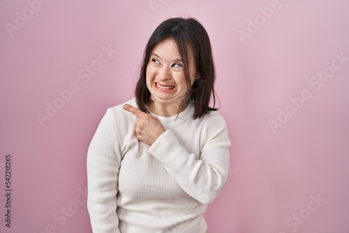 Woman with down syndrome standing over pink background pointing aside worried and nervous with forefinger  concerned and surprised expression