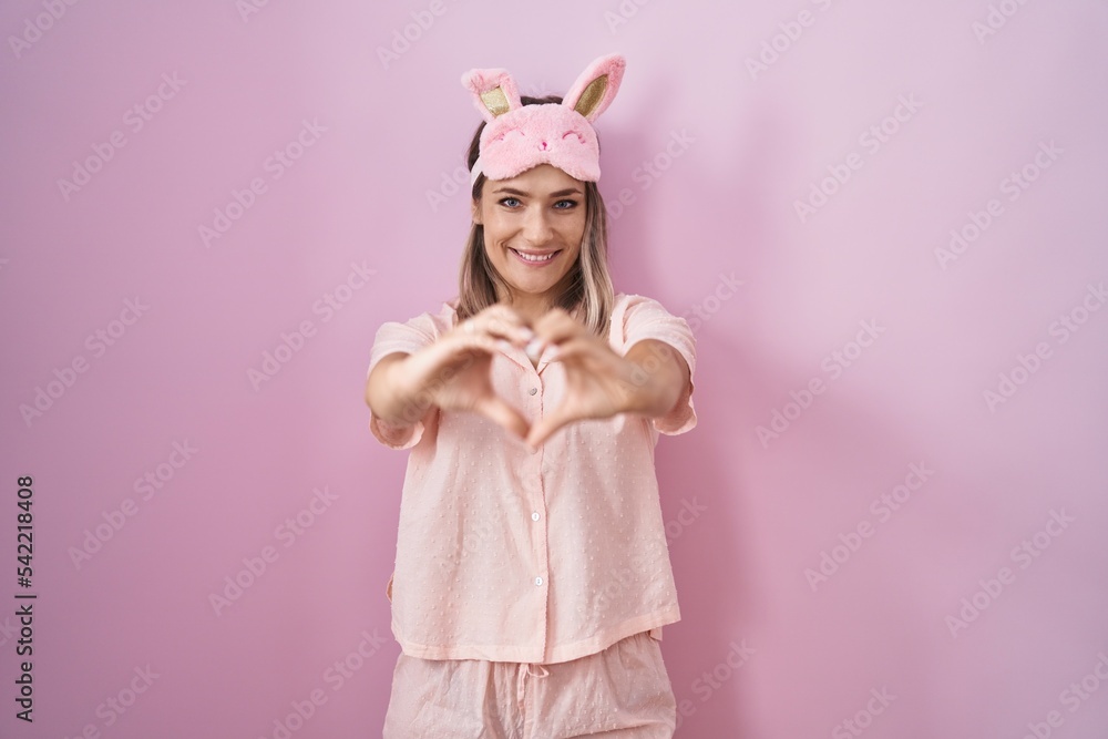 Blonde caucasian woman wearing sleep mask and pajama smiling in love doing heart symbol shape with hands. romantic concept.