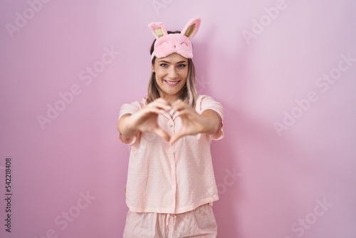 Blonde caucasian woman wearing sleep mask and pajama smiling in love doing heart symbol shape with hands. romantic concept.
