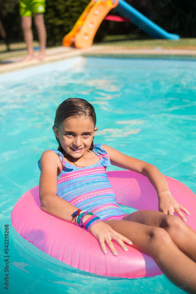 Close-up of smiling girl posing on inflatable ring in pool. Little fair-haired girl in blue swimsuit lying on big pink ring in water and looking at camera. Leisure activity and happy childhood concept