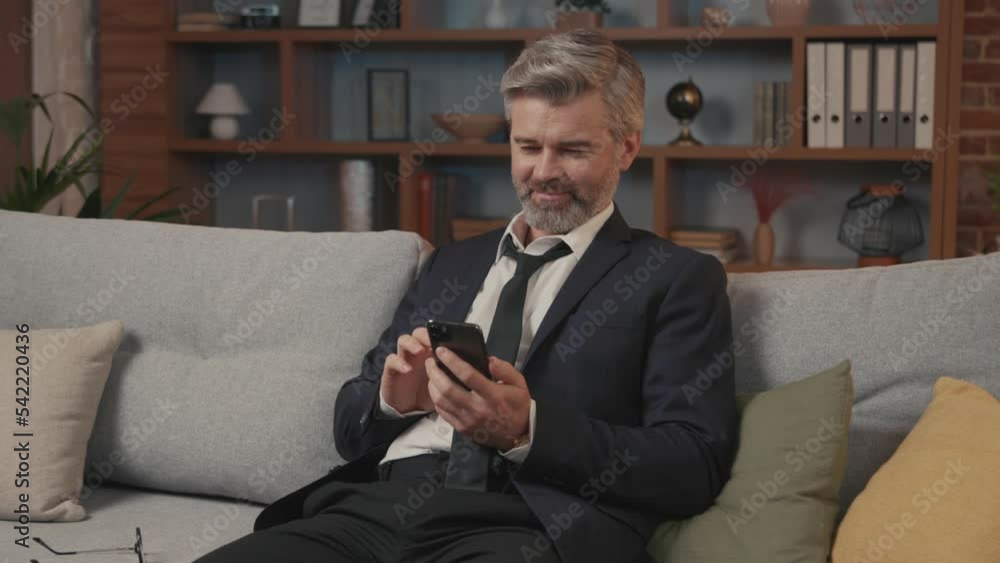 Portrait of stressed overwhelmed exhausted businessman in suits sits on the couch use phone and feel tired. Come back to home after hard day work. Modern apartment. Slow motion Portrait of stressed overwhelmed exhausted businessman in suits sits on the couch use phone and feel tired. Come back to home after hard day work. Modern apartment. Slow motion