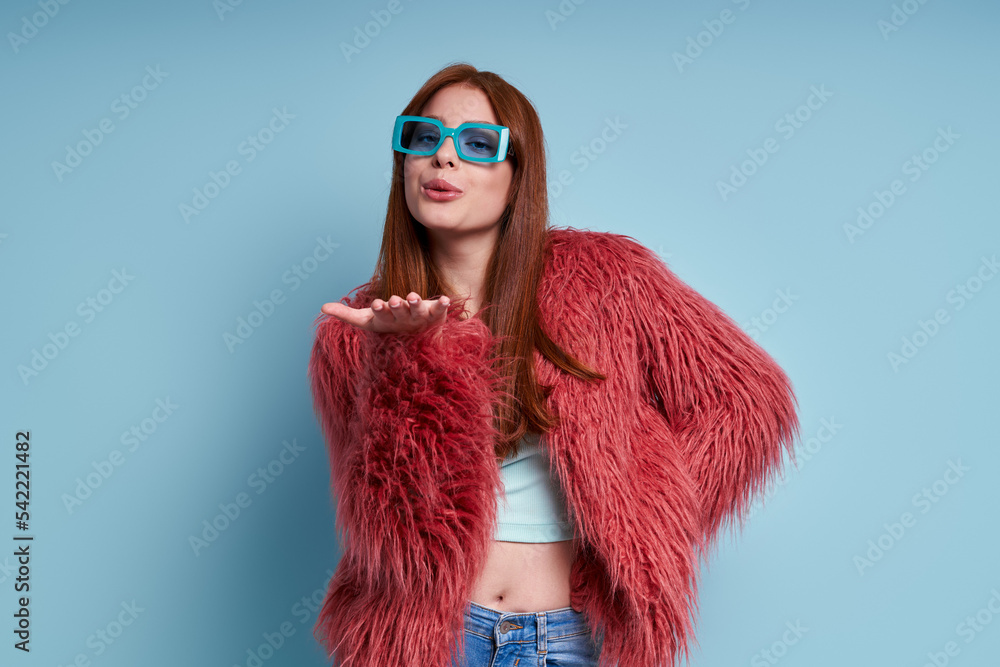 Happy young woman in fluffy coat blowing a kiss while standing against blue background Happy young woman in fluffy coat blowing a kiss while standing against blue background