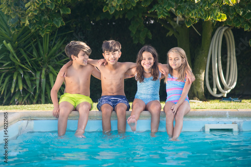 Portrait of happy Caucasian children wetting their legs in pool. Two boys and two girls sitting on edge of pool with legs in blue water laughing and hugging each other. Leisure and friendship concept