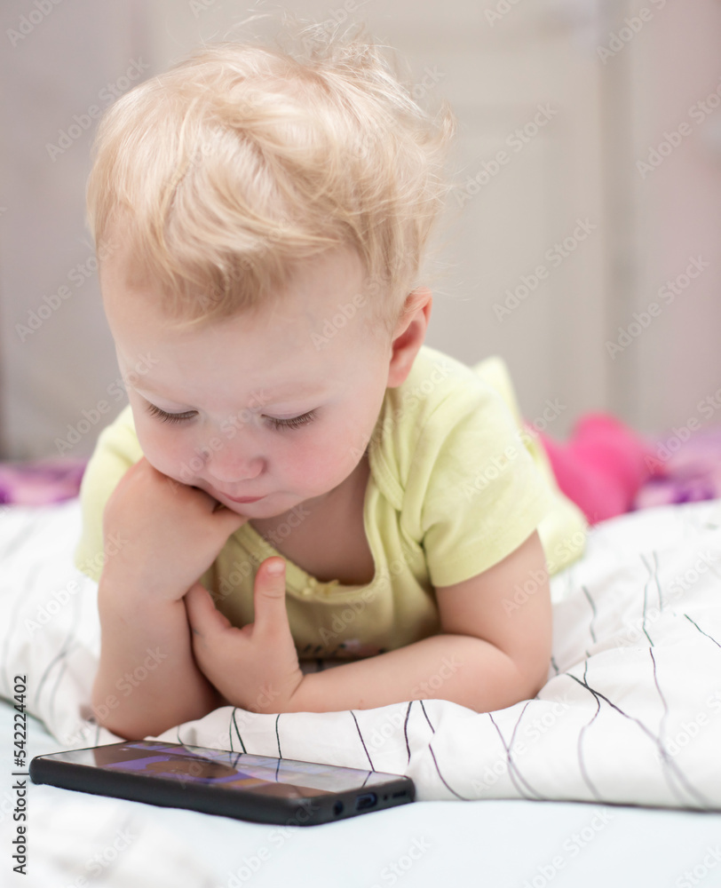 small child watches cartoons on mobile phone lying on bed. girl using smartphone to play at home. child scratches cutting teeth with finger. Stay home concept. Prevention of coronavirus infection small child watches cartoons on mobile phone lying on bed. girl using smartphone to play at home. child scratches cutting teeth with finger. Stay home concept. Prevention of coronavirus infection